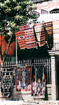 Colorful, patterned textiles hang over a metal fence in an outdoor setting. Several rugs are displayed, showcasing intricate designs and vibrant colors. A nearby tree with green leaves provides partial shade, and a stone building with textured walls stands in the background.