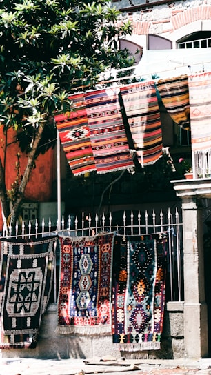 Colorful, patterned textiles hang over a metal fence in an outdoor setting. Several rugs are displayed, showcasing intricate designs and vibrant colors. A nearby tree with green leaves provides partial shade, and a stone building with textured walls stands in the background.