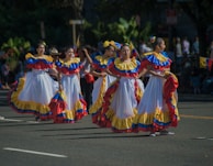 A group of people are participating in a parade, wearing traditional, vibrant dresses with ruffles in red, blue, and yellow. The scene is lively and festive, with participants smiling and walking in formation. Trees and a small crowd are visible in the background, suggesting an outdoor environment.