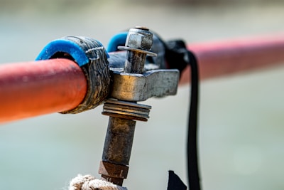 Close-up of a metal clamp securing a red rod, with visible blue padding for protection and a rope tied around it. The background is blurred, creating a shallow depth of field effect.