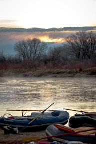 Inflatable boats are resting on a sandy riverbank in front of a calm river. The early morning or late evening sky is filled with soft clouds and the sun is setting or rising behind a line of bare trees across the water. The peaceful scene is enhanced by the stillness of the water reflecting the subtle light.