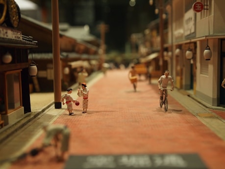 A miniature diorama depicts a street scene, featuring small-scale models of people in traditional clothing, a cyclist, and various buildings. The figures are engaged in activities such as walking and cycling along a brick-paved street lined with lanterns.