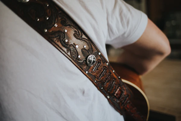 A tooled-leather guitar strap with brass conchos, worn over a white t-shirt &mdash; the kind a working road musician wears down over a decade.