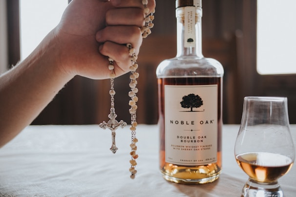 A hand is holding a rosary with a cross pendant next to a bottle of Noble Oak bourbon whiskey. A glass of whiskey partially filled sits beside the bottle on a table with a soft focus background.