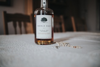 A bottle of Noble Oak Double Oak Bourbon stands on a textured white tablecloth. The bottle features a label with a stylized tree logo and descriptive text. In the background, wooden chairs are softly out of focus, creating a warm, inviting atmosphere. A small assortment of scattered seeds or grains is present on the table.
