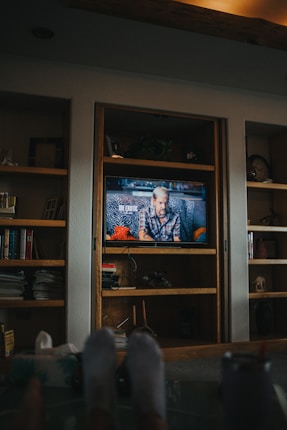 A cozy living room setting featuring a wooden shelf unit with a television at the center. The TV displays a man sitting on a sofa with zebra-patterned cushions. Surrounding the TV are various books and decorative items. Two out-of-focus feet in socks rest in the foreground. Soft, warm lighting creates a relaxed atmosphere.