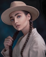 A woman with braided hair wears a wide-brimmed hat and a light-colored jacket. Her makeup is natural, highlighting her eyes and lips, and she gazes directly at the viewer. There is a subtle tribal-inspired pattern on her forehead. The background is dark and out of focus, emphasizing her poised expression.