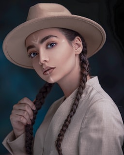 A woman with braided hair wears a wide-brimmed hat and a light-colored jacket. Her makeup is natural, highlighting her eyes and lips, and she gazes directly at the viewer. There is a subtle tribal-inspired pattern on her forehead. The background is dark and out of focus, emphasizing her poised expression.