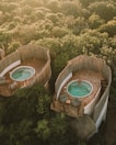 Aerial view of two rounded, rustic structures nestled in lush greenery, each featuring a private patio with a small pool. One of the pools is occupied by a person relaxing in the water, while the patios are furnished with comfortable seating and small tables. The surrounding area is heavily wooded, providing a serene and natural setting.