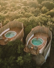 Aerial view of two rounded, rustic structures nestled in lush greenery, each featuring a private patio with a small pool. One of the pools is occupied by a person relaxing in the water, while the patios are furnished with comfortable seating and small tables. The surrounding area is heavily wooded, providing a serene and natural setting.
