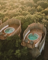 Aerial view of two rounded, rustic structures nestled in lush greenery, each featuring a private patio with a small pool. One of the pools is occupied by a person relaxing in the water, while the patios are furnished with comfortable seating and small tables. The surrounding area is heavily wooded, providing a serene and natural setting.