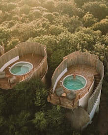 Aerial view of two rounded, rustic structures nestled in lush greenery, each featuring a private patio with a small pool. One of the pools is occupied by a person relaxing in the water, while the patios are furnished with comfortable seating and small tables. The surrounding area is heavily wooded, providing a serene and natural setting.