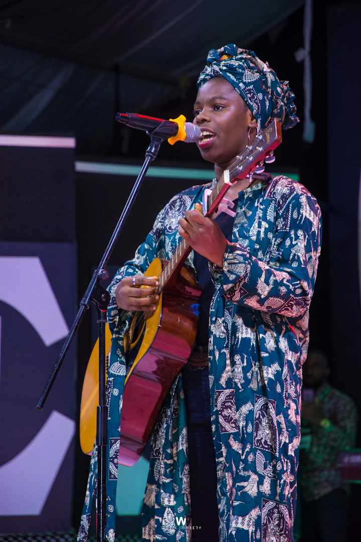 Une chanteuse en ensemble et foulard imprimés, guitare en bandoulière, en train de chanter devant un micro sur scène.