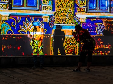 A vibrant scene where bright and colorful patterns are projected on a building facade. One person stands in front of the wall, posing, while another person is crouched down, seemingly taking a photo of the first person. Both their shadows are cast against the wall, becoming part of the intricate light display.
