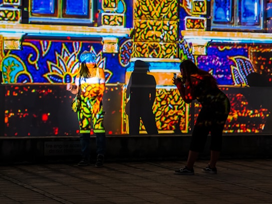 A vibrant scene where bright and colorful patterns are projected on a building facade. One person stands in front of the wall, posing, while another person is crouched down, seemingly taking a photo of the first person. Both their shadows are cast against the wall, becoming part of the intricate light display.