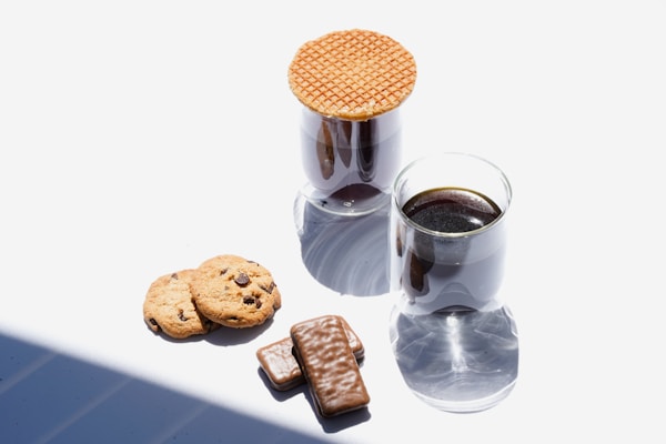 Two glasses of coffee are placed on a white surface, one topped with a waffle cookie. Nearby, two chocolate chip cookies and two chocolate-covered rectangular biscuits are arranged.