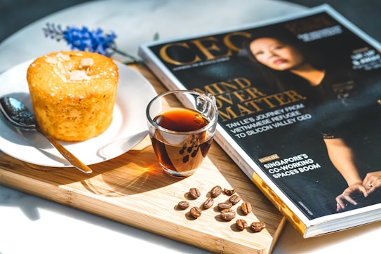 A wooden tray holds a glossy magazine featuring a cover photo and headlines. Next to it, a glass of dark coffee sits beside a small round cake topped with almond slices on a white plate. Coffee beans are scattered on the tray, and there's a silver spoon resting next to the cake. A small bunch of purple flowers lies in the background.