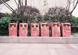 Six red fire hydrant valves are aligned on a paved sidewalk, situated in front of neatly trimmed bushes and a building facade. Each valve has a distinctive pair of circular outlets, giving them an intriguing, owl-like appearance. Metal plaques with inscriptions are attached to the front of each valve.