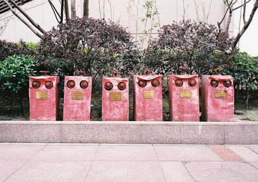 Six red fire hydrant valves are aligned on a paved sidewalk, situated in front of neatly trimmed bushes and a building facade. Each valve has a distinctive pair of circular outlets, giving them an intriguing, owl-like appearance. Metal plaques with inscriptions are attached to the front of each valve.