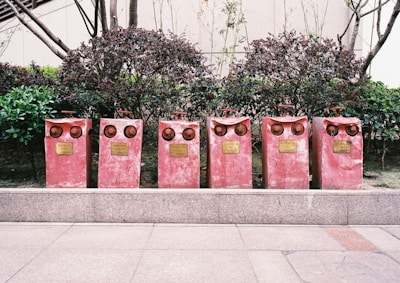Six red fire hydrant valves are aligned on a paved sidewalk, situated in front of neatly trimmed bushes and a building facade. Each valve has a distinctive pair of circular outlets, giving them an intriguing, owl-like appearance. Metal plaques with inscriptions are attached to the front of each valve.