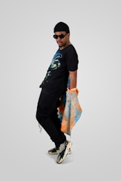 A person wearing a black beanie, sunglasses, and a graphic T-shirt is posed in a confident stance. They hold an orange and blue patterned cloth draped over their arm. The person is wearing black pants and stylish sneakers.