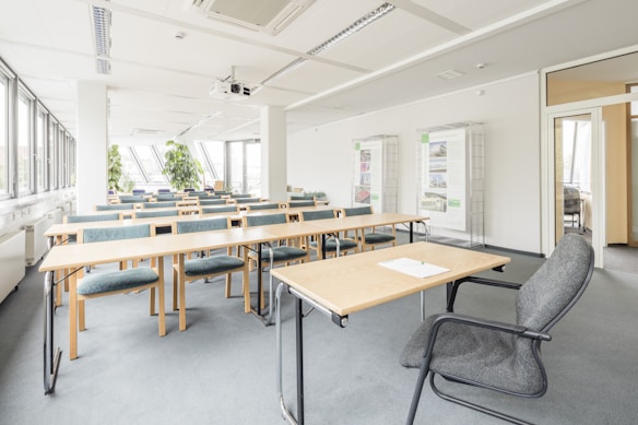 A bright, empty classroom with rows of wooden desks and upholstered chairs arranged neatly. Large windows on the left side allow natural light to fill the space, creating a clean and airy atmosphere. Green plants are placed near the windows for decoration. The room is equipped with a projector mounted on the ceiling and displays in the background.