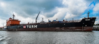 A large cargo ship is docked in a port area with a body of water in the foreground. The ship is painted in red and black colors, with the name 'TORM' and another name, 'TORM OHIO', visible on its side. The ship has multiple cranes and structures on its deck. The sky above is mostly cloudy with some patches of blue.