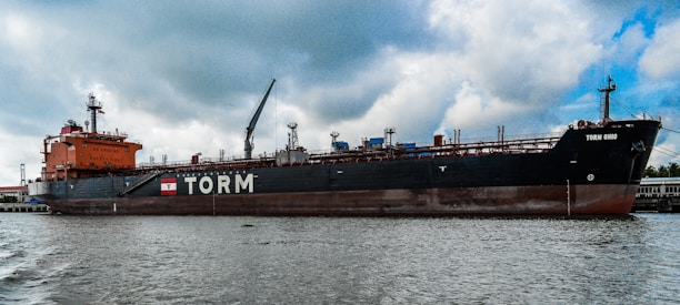 A large cargo ship is docked in a port area with a body of water in the foreground. The ship is painted in red and black colors, with the name 'TORM' and another name, 'TORM OHIO', visible on its side. The ship has multiple cranes and structures on its deck. The sky above is mostly cloudy with some patches of blue.