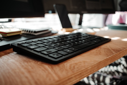 A close-up view of a black keyboard on a wooden desk, accompanied by office supplies such as stacked notebooks and sticky notes in the background. A blurred monitor stands at the back, creating a workspace vibe.