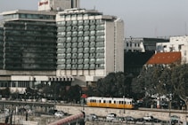 A multi-story hotel building with a modern architectural design is situated near a street with trees lining the sidewalk. A yellow tram is seen moving along the road in front of parked cars. The sky is clear, and several smaller buildings are visible in the background.
