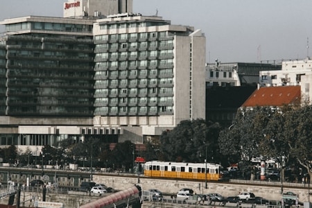 A multi-story hotel building with a modern architectural design is situated near a street with trees lining the sidewalk. A yellow tram is seen moving along the road in front of parked cars. The sky is clear, and several smaller buildings are visible in the background.
