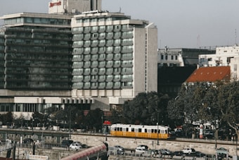 A multi-story hotel building with a modern architectural design is situated near a street with trees lining the sidewalk. A yellow tram is seen moving along the road in front of parked cars. The sky is clear, and several smaller buildings are visible in the background.
