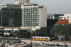 A multi-story hotel building with a modern architectural design is situated near a street with trees lining the sidewalk. A yellow tram is seen moving along the road in front of parked cars. The sky is clear, and several smaller buildings are visible in the background.