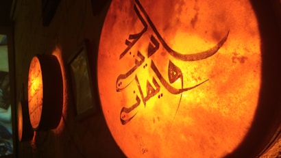 A dimly lit wall features circular objects with intricate Arabic calligraphy illuminated from behind, creating a warm, ambient glow. The composition highlights the elegant curves and lines of the script, enhancing its artistic appeal.