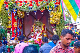 A vibrant and colorful celebration with a decorated platform featuring floral garlands, fruits, and a central statue adorned with various offerings. People are gathered around, creating a lively and festive atmosphere.