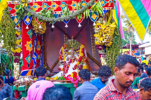 A vibrant and colorful celebration with a decorated platform featuring floral garlands, fruits, and a central statue adorned with various offerings. People are gathered around, creating a lively and festive atmosphere.