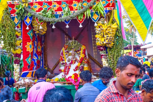 A vibrant and colorful celebration with a decorated platform featuring floral garlands, fruits, and a central statue adorned with various offerings. People are gathered around, creating a lively and festive atmosphere.
