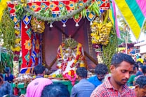A vibrant and colorful celebration with a decorated platform featuring floral garlands, fruits, and a central statue adorned with various offerings. People are gathered around, creating a lively and festive atmosphere.