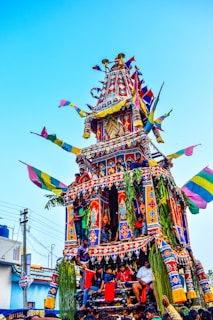 A vibrant, multi-tiered chariot adorned with colorful flags and intricate patterns is being pushed through the street by a group of people. The chariot is decorated with lush green leaves and ornate details, and there are people riding on different levels. Overhead, bright blue skies enhance the vivid colors of the scene.