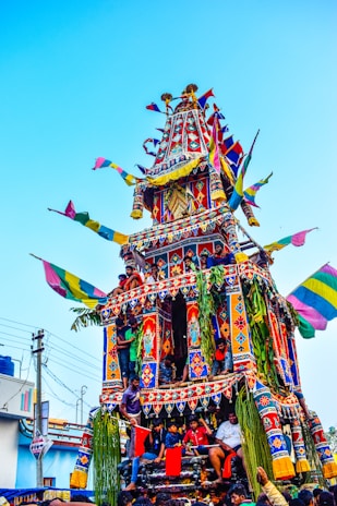 A vibrant, multi-tiered chariot adorned with colorful flags and intricate patterns is being pushed through the street by a group of people. The chariot is decorated with lush green leaves and ornate details, and there are people riding on different levels. Overhead, bright blue skies enhance the vivid colors of the scene.