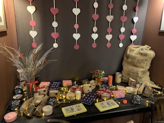 A table is adorned with decorative gift boxes, candles, and trinkets. Above, heart and circle cutouts hang on strings against a dark backdrop. To the left, a vase holds dry grasses with red accents, and a large burlap sack is situated on the right. The table features warm string lights, enhancing a cozy ambiance.