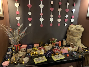 A table is adorned with decorative gift boxes, candles, and trinkets. Above, heart and circle cutouts hang on strings against a dark backdrop. To the left, a vase holds dry grasses with red accents, and a large burlap sack is situated on the right. The table features warm string lights, enhancing a cozy ambiance.