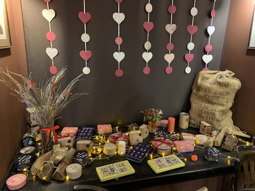A table is adorned with decorative gift boxes, candles, and trinkets. Above, heart and circle cutouts hang on strings against a dark backdrop. To the left, a vase holds dry grasses with red accents, and a large burlap sack is situated on the right. The table features warm string lights, enhancing a cozy ambiance.