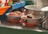 Bowls filled with vibrant noodle dishes rest on an orange plate, featuring finely chopped vegetables and a crispy element on top. A large metal pot emits steam in the background, suggesting hot food preparation. Bottles of sauce and utensils are visible, contributing to a busy kitchen setting.