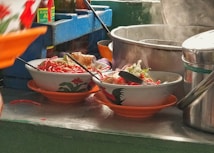 Bowls filled with vibrant noodle dishes rest on an orange plate, featuring finely chopped vegetables and a crispy element on top. A large metal pot emits steam in the background, suggesting hot food preparation. Bottles of sauce and utensils are visible, contributing to a busy kitchen setting.