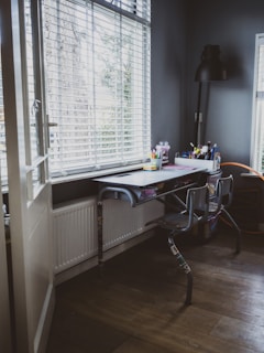 A cozy workspace with a metal desk and two chairs is situated by large windows with blinds, allowing soft natural light to illuminate the room. Various stationery supplies, such as pens and notebooks, are organized neatly on the desk. A tall black desk lamp stands in the corner, providing additional light if needed. The walls and floor are in dark tones, creating a calm and focused atmosphere.