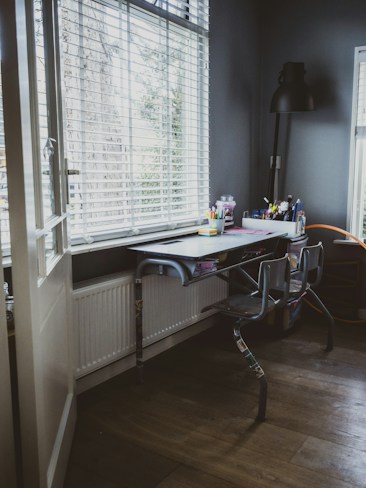 A cozy workspace with a metal desk and two chairs is situated by large windows with blinds, allowing soft natural light to illuminate the room. Various stationery supplies, such as pens and notebooks, are organized neatly on the desk. A tall black desk lamp stands in the corner, providing additional light if needed. The walls and floor are in dark tones, creating a calm and focused atmosphere.