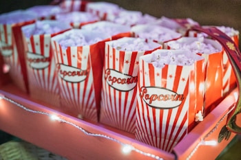 Multiple paper bags filled with popcorn are neatly arranged in a tray. The bags have a retro design with red and white vertical stripes and the word 'Popcorn' printed on them. Warm fairy lights are draped around, creating a festive and inviting atmosphere.