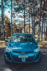 A blue Hyundai car is parked on a road surrounded by tall trees. The car is facing forward, and the scene is set in a forested area during the day. The shadows of the trees are reflected on the car's hood.
