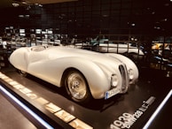 A vintage white sports car displayed in a dimly lit showroom with reflective black walls. The car has a sleek, aerodynamic body with a rounded front, prominent headlights, and a clearly visible license plate. Information and images about the car are placed alongside it on a panel.